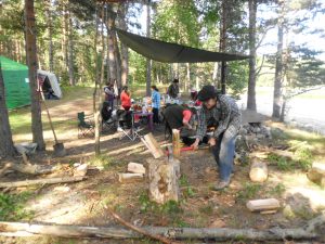 A man chops wood on a tree stump at a forest campsite, while several people gather around a table under a tarp. Tents are set up nearby and trees surround the area.