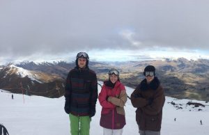 Three people in ski gear stand on a snowy mountain slope with arms crossed, smiling at the camera. Snow-covered peaks and a wide valley stretch into the distance under a cloudy sky.