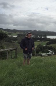 A person wearing a dark jacket, patterned shorts, and a beanie stands on green grass with hills, trees, and a body of water in the background under a cloudy sky.