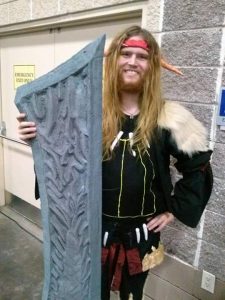 A person in costume with a long blond wig, elf ears, a red headband, and a black tunic with yellow lines poses indoors holding a large, textured, gray foam sword. They are smiling and standing against a gray brick wall.