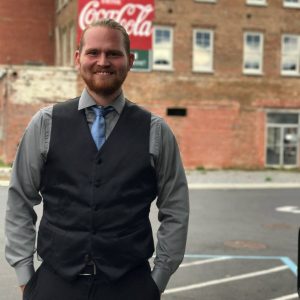 A smiling man with a beard, wearing a gray shirt, blue tie, and black vest, stands outdoors in front of a brick building with a Coca-Cola sign.