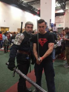 Two men at a convention pose together; one is dressed as Geralt from The Witcher with armor and sword, while the other wears a black Superman shirt and holds a sword. People and vendor booths are visible in the background.