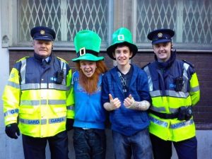 Two smiling young people in oversized green leprechaun hats pose with two uniformed Irish police officers (Gardaí) in high-visibility jackets, standing against a brick wall with barred windows.