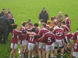 A group of young male athletes in maroon and white jerseys celebrate together on a grassy field, some hugging and smiling, while coaches and adults look on.