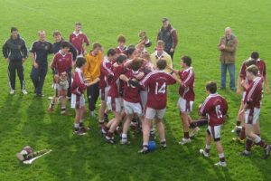 A group of young athletes in maroon and white uniforms celebrate on a grass field, some hugging and others clapping, while a few adults look on and a trophy is raised in the center of the group.