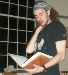 A man with long hair wearing a striped headband and black t-shirt stands indoors, holding and reading a large open book. He touches his neck with one hand and looks focused. A window is visible in the background.