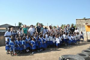 A large group of young children in matching blue and white uniforms stand in front of a group of adults outdoors, posing for a photo on a sunny day, with buildings and clear blue sky in the background.