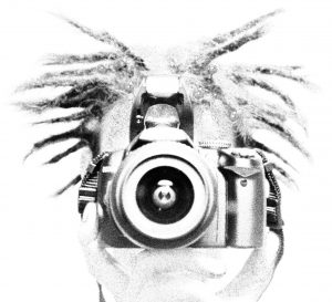 Black and white, high-contrast image of a person with spiky hair holding a camera up to their face, as if taking a picture. The focus is on the camera lens, centered in the frame.