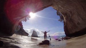 Two people stand with arms outstretched on a rock beneath a large cave arch on a sunny beach, with the bright sun shining through the opening and several people and rock formations in the background.