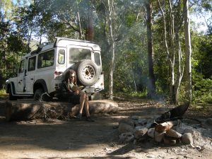 A person sits on a log reading near a campfire in a forest, with a white off-road vehicle parked nearby. Sunlight filters through the trees and smoke rises from the fire.