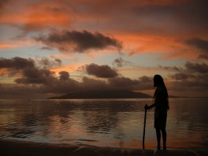 A person with long hair stands on a beach at sunset, holding a stick, silhouetted against colorful clouds and calm water with an island in the distance.
