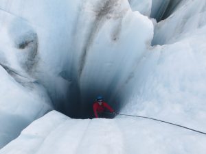 A climber in a red jacket and helmet ascends an icy crevasse using a rope, surrounded by towering walls of blue and white ice.