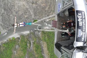 A person bungee jumps from the Nevis Highwire platform, 134 meters above a rocky canyon with a river below, as two people watch from the platform.