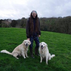 A person with long dreadlocks, wearing a headband, jacket, jeans, and boots, stands on grass holding leashes of two smiling golden retrievers, with trees and a cloudy sky in the background.