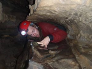 A person wearing a red helmet with a headlamp crawls through a narrow, rocky cave passage. The surroundings are damp and the space is tight, requiring careful maneuvering.