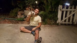A man sits on a concrete patio at night, smiling while holding a cat. He wears a light yellow t-shirt, dark shorts, and sneakers. There are plants, bricks, and a white picket gate in the background.