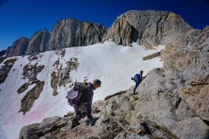 Two climbers with backpacks and helmets trek across a rocky, snowy mountain ridge under a deep blue sky, with jagged peaks rising in the background.