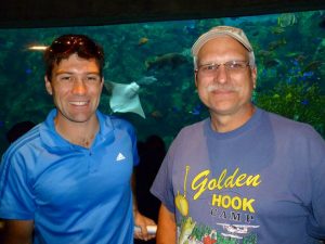 Two men smiling in front of a large aquarium tank filled with fish and a stingray. One man wears a blue shirt and sunglasses on his head; the other wears glasses, a cap, and a Golden Hook Camp T-shirt.