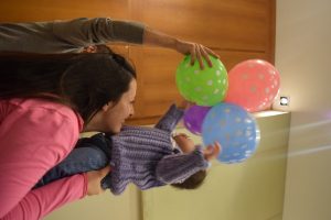 An adult lifts a small child wearing a purple sweater toward colorful polka-dot balloons while another person holds the balloons up. The scene appears joyful and playful indoors.