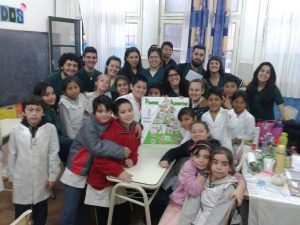 A group of children and adults gather in a classroom around a table displaying a colorful project poster. The students wear white coats and smile at the camera, with classroom supplies visible around them.