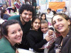 A group of two adults and three children smile for a selfie in a colorful classroom filled with students and posters in the background.