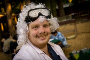 A smiling person with a white curly wig and black goggles on their head wears a white lab coat, resembling a scientist, while outdoors with people and trees in the background.