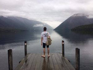 A person stands barefoot on a wooden dock facing a calm lake surrounded by misty mountains, holding a bag and wearing a white shirt that reads “NO TALK SUMMER.” The sky is overcast.