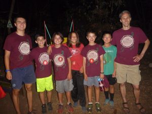 Seven people, including Randall Grayson, stand in a row outdoors at night—six boys and one adult. Most wear matching maroon Camp Augusta shirts, while one wears red and another green. They smile brightly, appearing to enjoy a lively camp event.