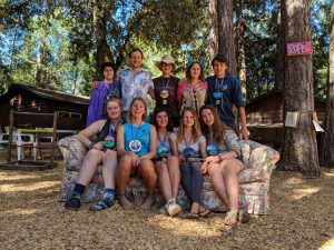 Nine people, including a summer camp counselor, pose for a group photo outdoors on a floral couch with trees and cabins in the background. Most wear camp medallions and casual clothes; a ROPES sign is on a tree to the right.