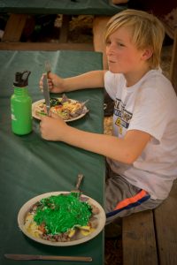 A blonde boy sits at a picnic table with a fork and knife, looking to the side. In front of him is a plate of food topped with bright green sauce, a water bottle, and another plate of food on the table.