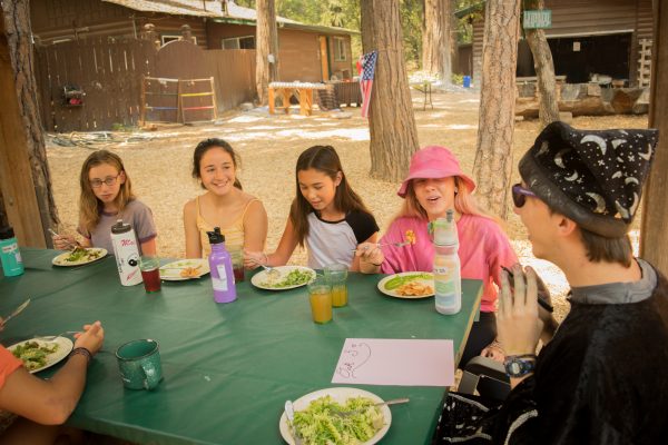 A group of kids and one adult in a wizard hat sit around a green table outdoors, eating a meal together and talking. Trees, wooden cabins, and an American flag are visible in the background.