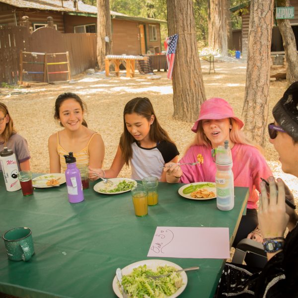 A group of kids and one adult in a wizard hat sit around a green table outdoors, eating a meal together and talking. Trees, wooden cabins, and an American flag are visible in the background.