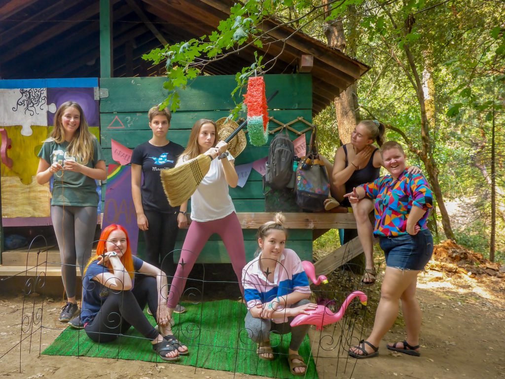 Seven young people pose playfully in front of a rustic cabin surrounded by trees; some hold props like a broom and pink flamingo, while others smile or make funny faces, creating a fun, casual atmosphere.