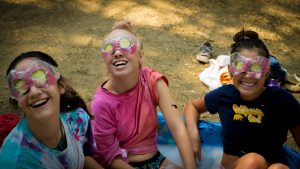 Three girls sitting together outdoors, smiling and laughing, each with a facial mask and cucumber slices over their eyes. They are enjoying a playful spa activity, with soap suds and casual clothes visible.