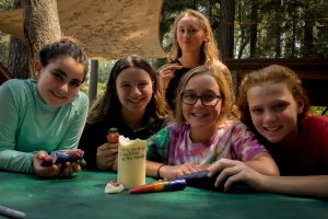 Five smiling girls sit at a green table outdoors, holding colorful candles and crafts. They are surrounded by trees and sunlight, suggesting a camp or nature setting. One girl in the back holds a paintbrush.