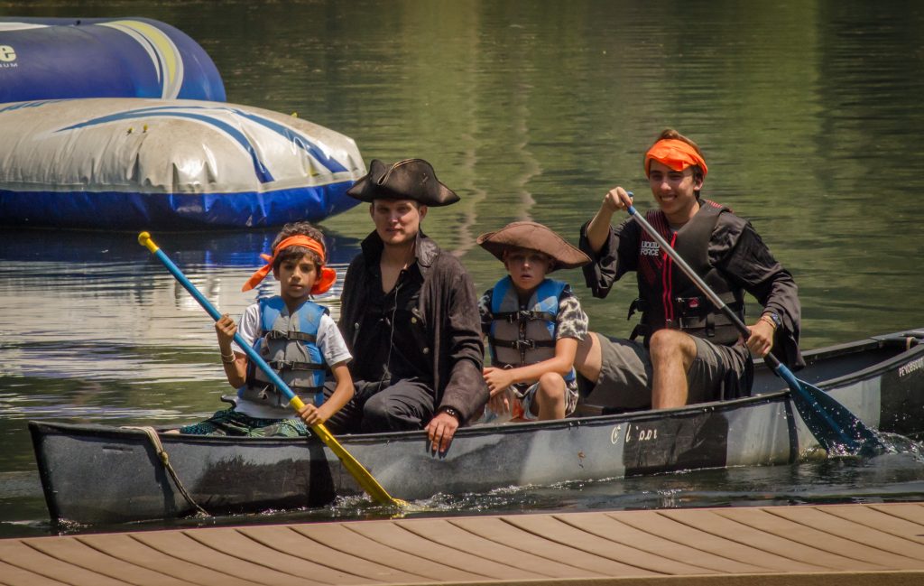 Four people, including two children and two adults dressed as pirates with hats and orange bandanas, paddle a canoe on a calm lake near a dock. An inflatable water structure floats in the background.