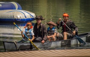 Four people, including two children and two adults dressed as pirates with hats and orange bandanas, paddle a canoe on a calm lake near a dock. An inflatable water structure floats in the background.