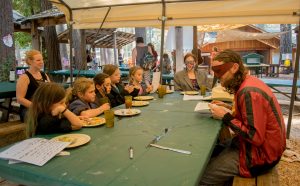 A group of children and two adults sit at a picnic table outdoors under a canopy, reading papers. One adult wears a red costume and mask; the other has face paint. Plates and cups are on the table, and cabins and trees are in the background.