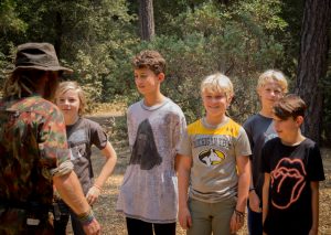 A group of five boys stands outdoors in a wooded area, smiling at a man in camouflage clothing and a hat who faces them. Sunlight filters through the trees, creating a warm atmosphere.