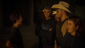 Four people stand together outside at night, smiling and talking. One person wears a cowboy hat and glasses; the others are dressed casually. Warm lighting illuminates their faces against a dark, blurred background.