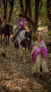 A woman in a light dress and boots leads a horse on a wooded trail. A girl in a helmet and purple shirt rides the horse, followed by another rider on a brown horse. The ground is covered with dry leaves.