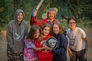 Six smiling kids pose outdoors; one holds a mask, another wears a toy helmet and wields a foam sword. The group is dressed in casual, comfy clothing, surrounded by trees and warm afternoon light.