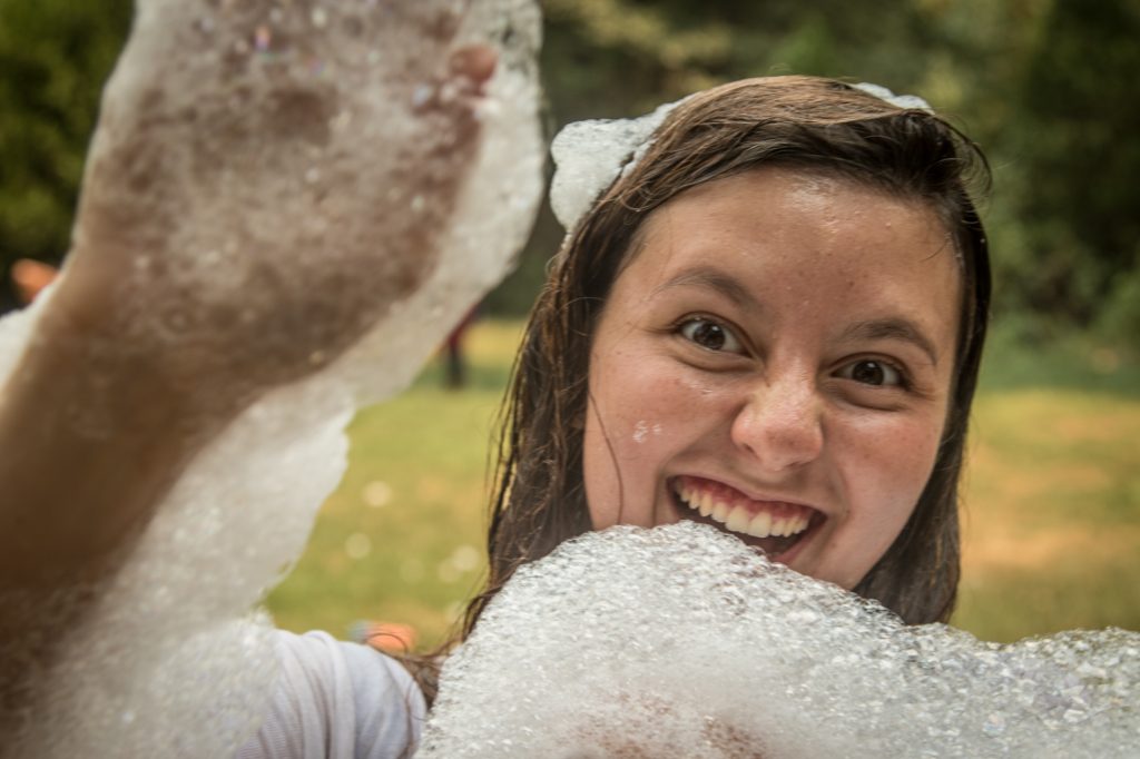 A young woman with wet hair smiles widely, surrounded by soap bubbles, holding up a foamy hand outdoors with green trees blurred in the background.