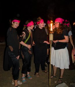 A group of people wearing black outfits and pink headbands stand together at night, smiling and talking near a lit torch. One person in a white skirt appears to be addressing the group.