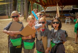 A group of children wearing sunglasses and gray shirts stand outdoors, smiling and holding clipboards. Some have green waist bags and whistles. One child speaks into a homemade megaphone. Trees and a wooden shelter are in the background.