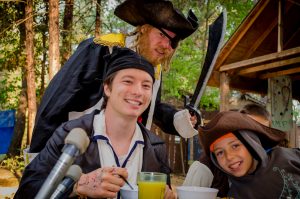 Three people dressed as pirates, two sitting and smiling with drinks, and one standing behind them with a sword, eye patch, and pirate hat, in an outdoor wooded area near a wooden structure.