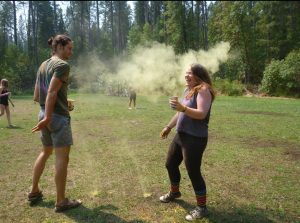 Two people stand on grass in a forested area; yellow powder covers the air between them. One person is laughing and holding a cup, while the other looks on. Trees and more people are visible in the background.