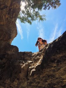 A woman wearing sunglasses smiles and leans over the edge of a rocky cliff, with bright blue sky and wispy clouds above. Sunlight filters through tree branches at the top of the image.