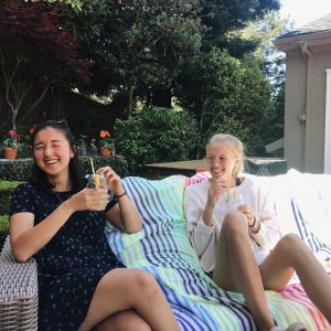 Two young women sit on a striped blanket outdoors, smiling and holding drinks with straws and lemon slices. They are surrounded by greenery and sunlight in what appears to be a backyard or garden setting.