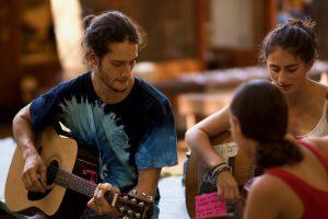 Three young adults sit together playing acoustic guitars. Two face forward, one in a tie-dye shirt, while the third person with a note on their guitar faces away. Warm, soft lighting creates a relaxed, informal atmosphere.